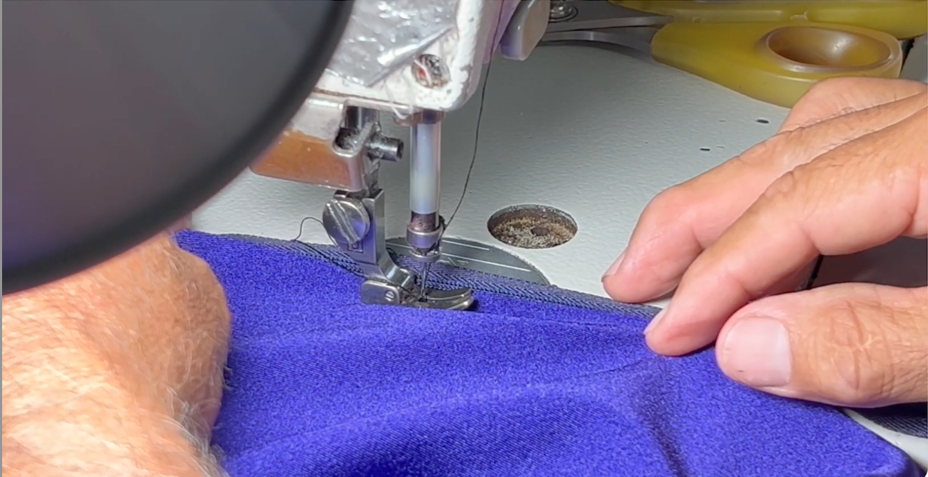 Hands guiding purple fabric under an industrial sewing machine at Doxa Couture in Downey, California.