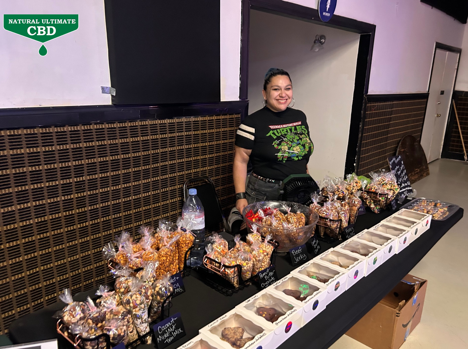 Vendor table with snacks and baked goods at Santino Bros Wrestling event in Azusa California with Natural Ultimate CBD logo visible